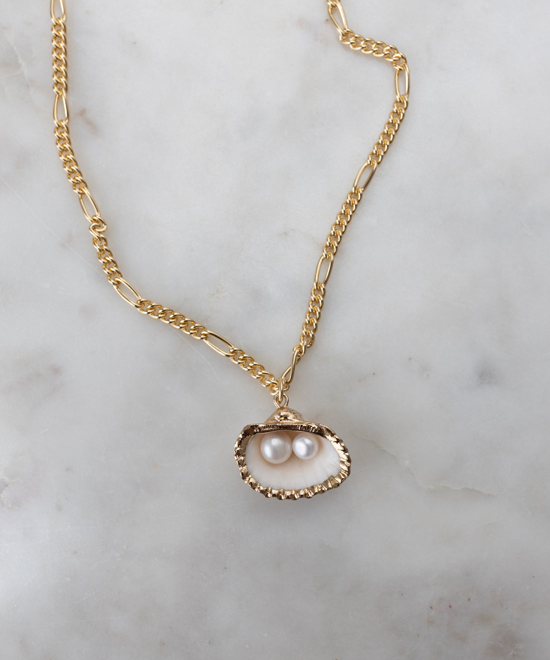 The Drop It Like It’s Hot Shell Necklace by WALD Berlin features an elegant gold-plated brass chain with an open oyster shell pendant and two pearls, shown handmade against a light marble background.