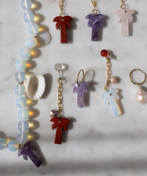 The Mexicana Mariachi Earrings Double Palm by WALD Berlin, along with beaded handmade jewelry, a shell, gold-plated accents, and a pearl ring are arranged on a light marble surface.