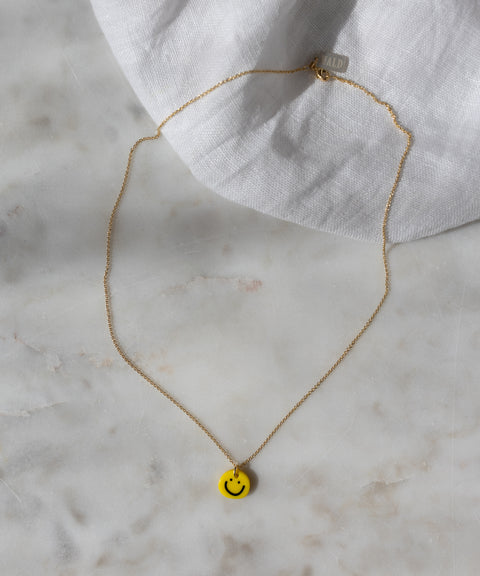 WALD Berlin’s Smilie Yellow Necklace, a gold-plated piece featuring a hand-painted pendant, is elegantly displayed on a marble surface with a white cloth in the background.