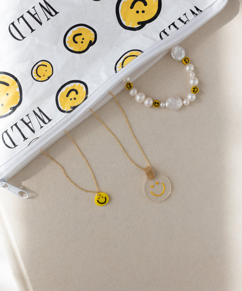 The WALD Berlin Smilie Yellow Necklace is arranged alongside a beaded bracelet and a clear hand-painted pendant with smiley faces, next to a white zipper pouch featuring a matching smiley design, all displayed on a beige surface.