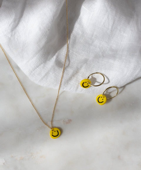 Smilie Yellow Earrings by WALD Berlin, featuring 18k gold-plated hoops with yellow smiley face charms, displayed on a white fabric and marble surface.