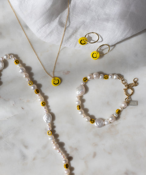 Eine Perlenkette und ein Armband, verziert mit gelben Smiley-Perlen, eine goldene Kette sowie die „Smilie Yellow“-Ohrringe von WALD Berlin – vergoldete Creolen mit passenden, in Deutschland gefertigten Smiley-Anhängern – präsentiert auf einer weißen Marmorfläche.