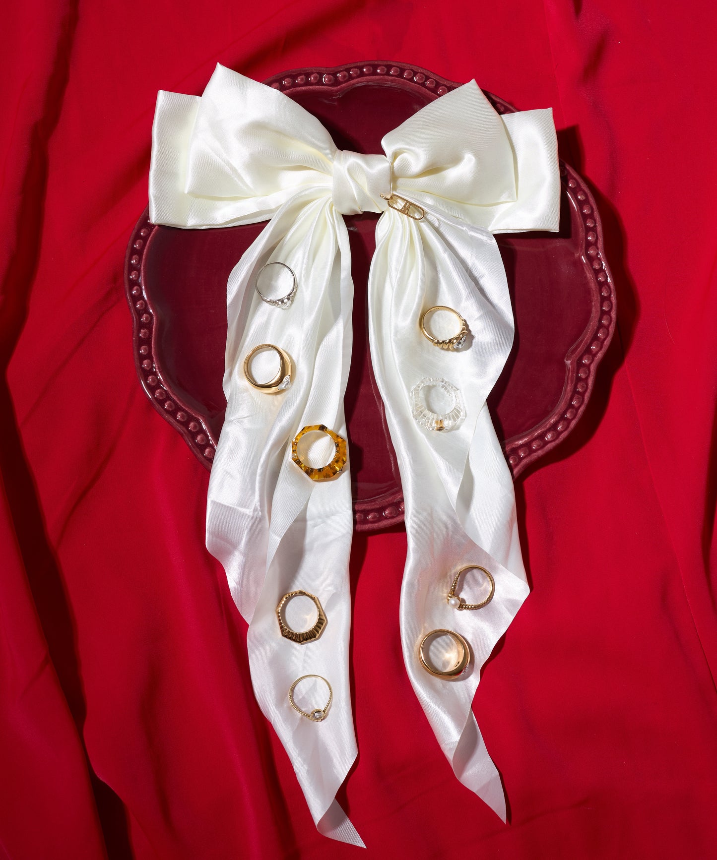 A white satin bow with eight stacked Shining Star Gold Rings by WALD Berlin in silver, gold, and gold-plated sterling silver sits on a red plate, set against rich red fabric.