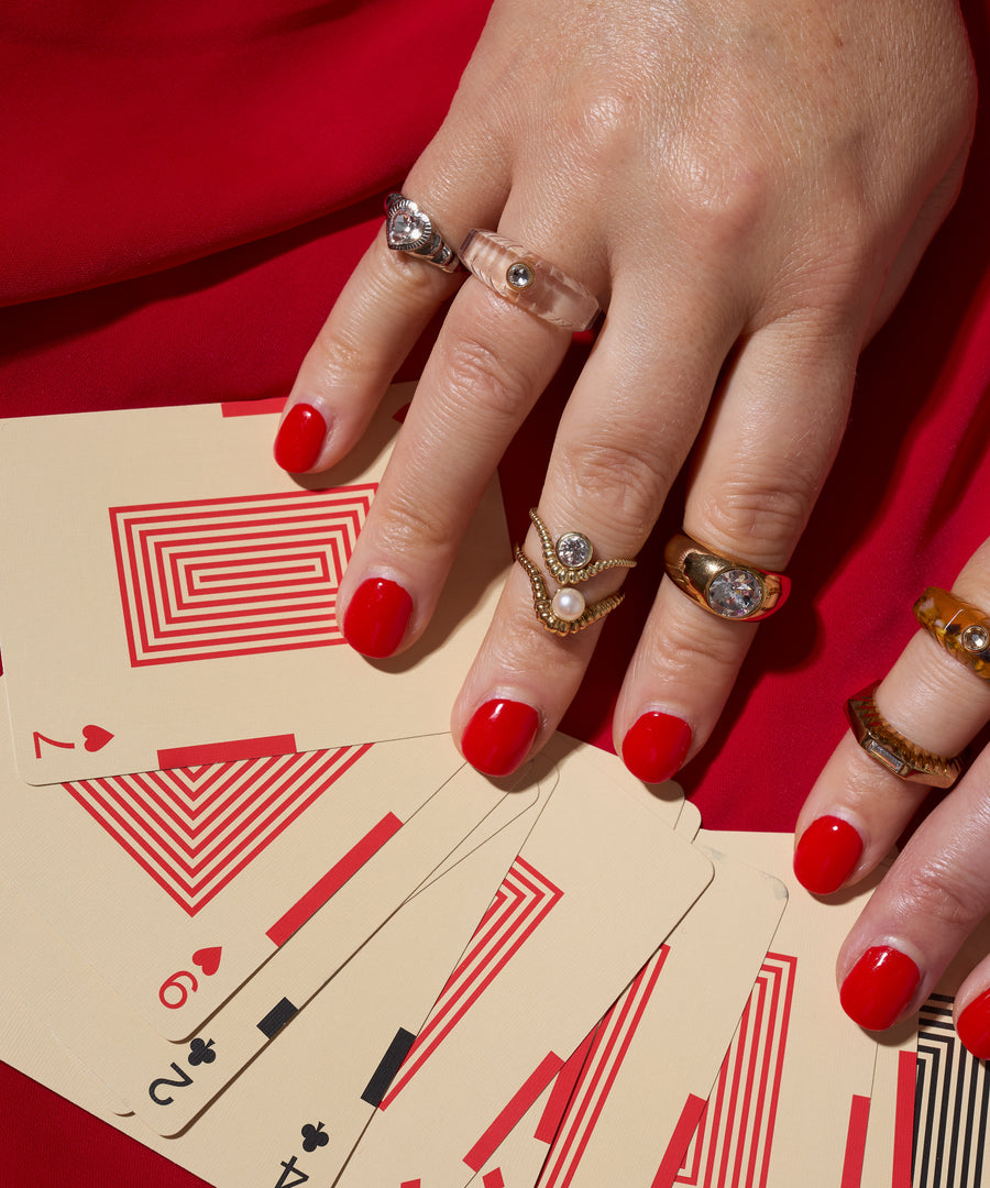 A hand with red nail polish wears the WALD Berlin NYC If I Rule The World Ring, resting on playing cards with geometric red designs on a matching surface.