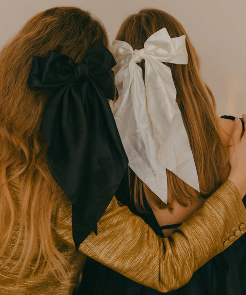 Two individuals with long hair, each highlighting a festive hair accessory—one featuring WALD Berlin's "Happy Holidays Black Hair Bow" and the other in white—bring a festive flair to their holiday hairstyles. The person on the left catches the eye with a shimmering gold top, ideal for the season's parties and gatherings.