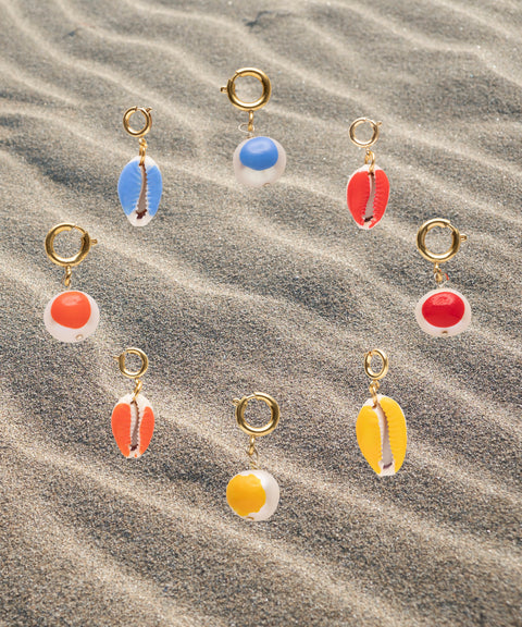 Nine gold-plated brass earrings from WALD Berlin's SAMPLE SALE Beach Babe Charms, featuring vibrant cowrie shells and circular pendants in blue, red, orange, and yellow, are artfully arranged on rippled sand.