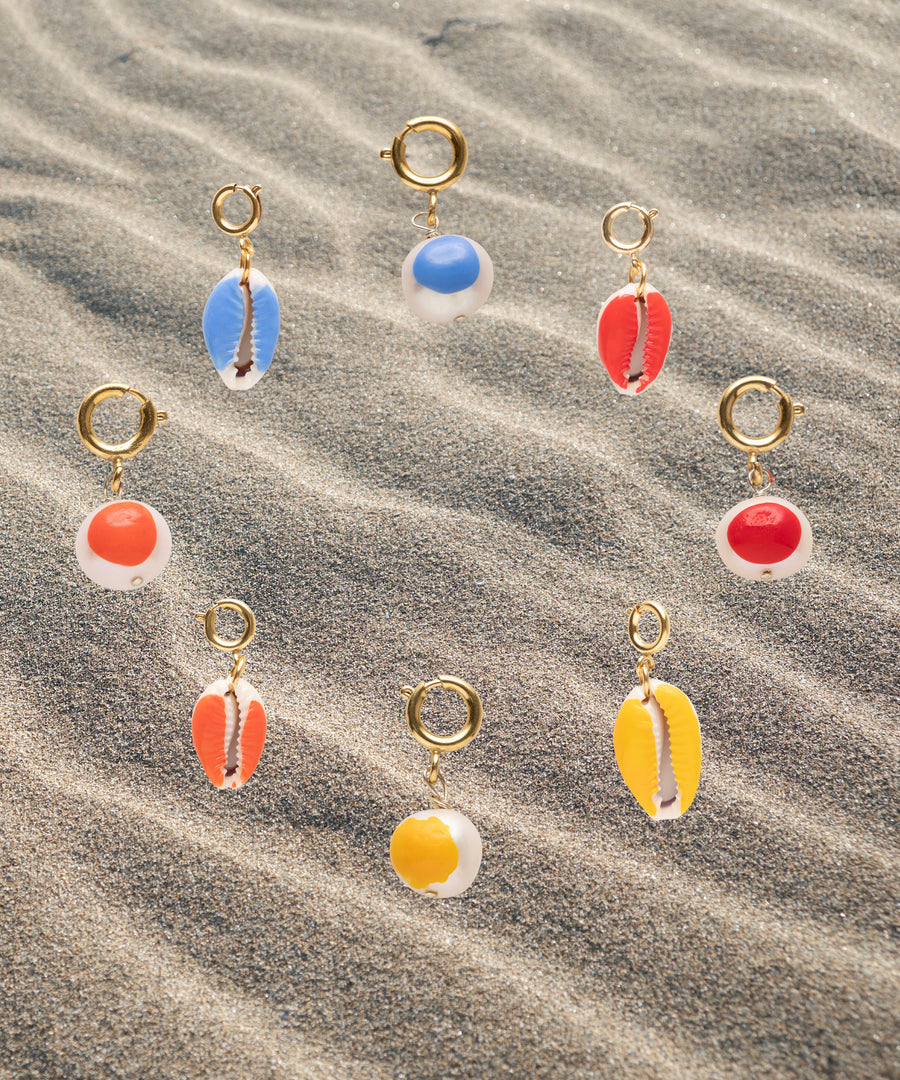 Nine gold-plated brass earrings from WALD Berlin's SAMPLE SALE Beach Babe Charms, featuring vibrant cowrie shells and circular pendants in blue, red, orange, and yellow, are artfully arranged on rippled sand.