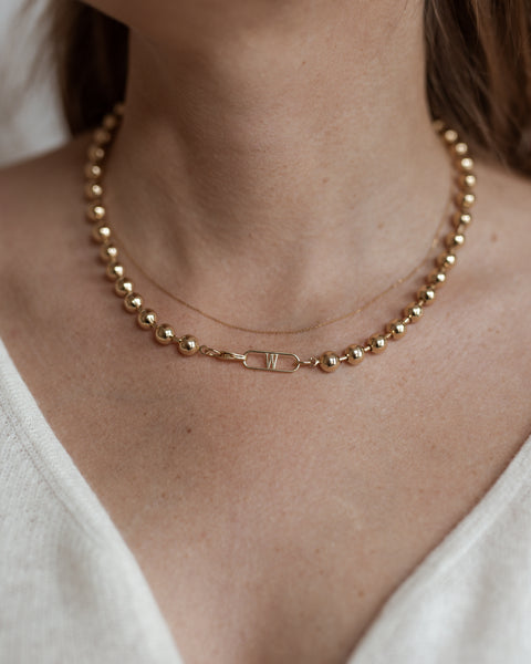 A close-up of a person wearing the Harry Necklace Gold by WALD Berlin, a gold-plated brass necklace featuring a small rectangular pendant with the letter "W," elegantly draped on a thin chain. The German jewelry piece beautifully complements their light-colored top.
