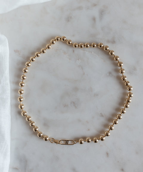 The WALD Berlin Harry Necklace Gold, a gold-plated brass beaded necklace featuring a clasp with the letter "W," is elegantly arranged on a marble surface.