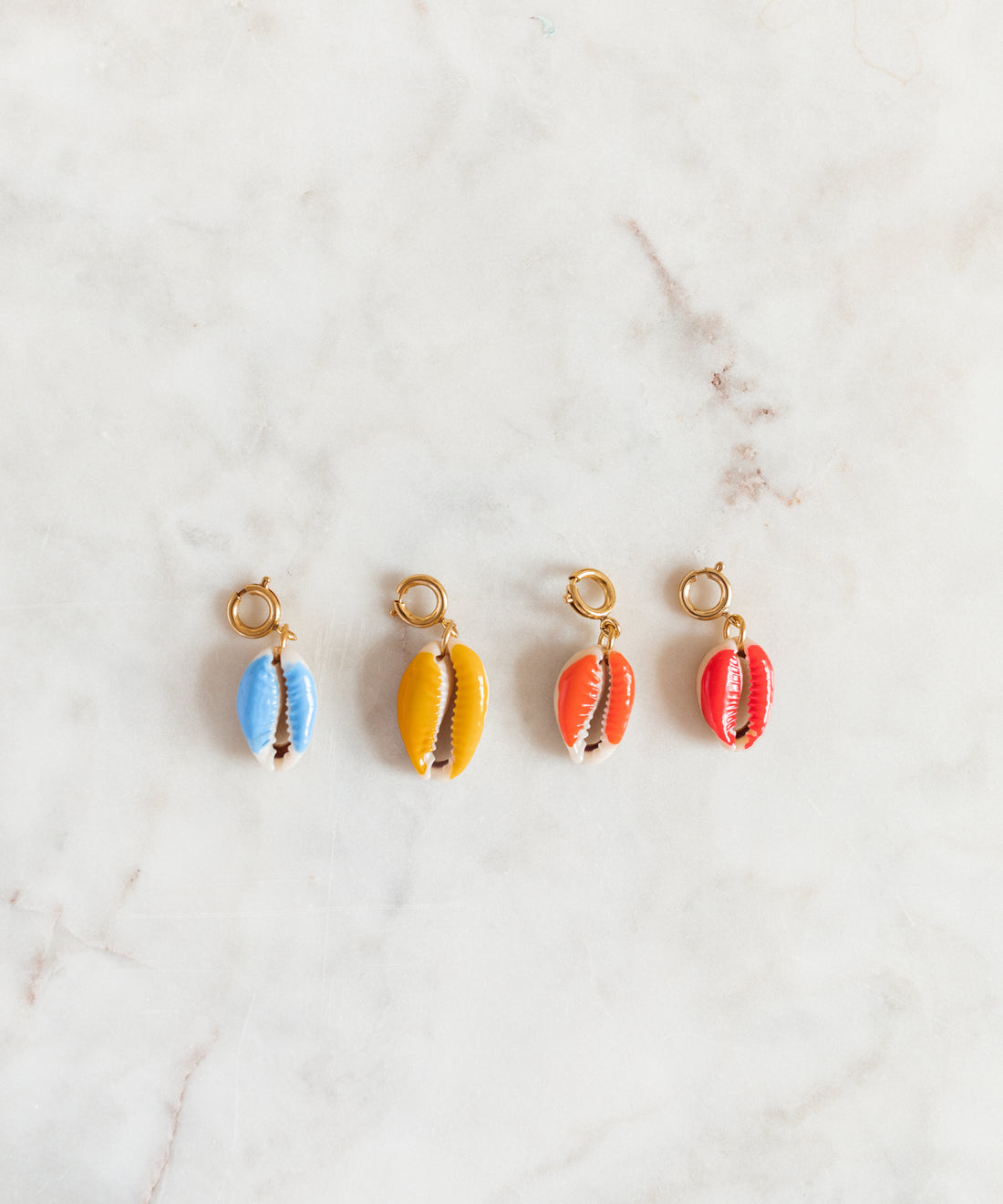 Four SAMPLE SALE Beach Babe Charms by WALD Berlin, each cowrie shell with a gold loop and painted in blue, yellow, orange, and red, are arranged in a row on a light marble surface.
