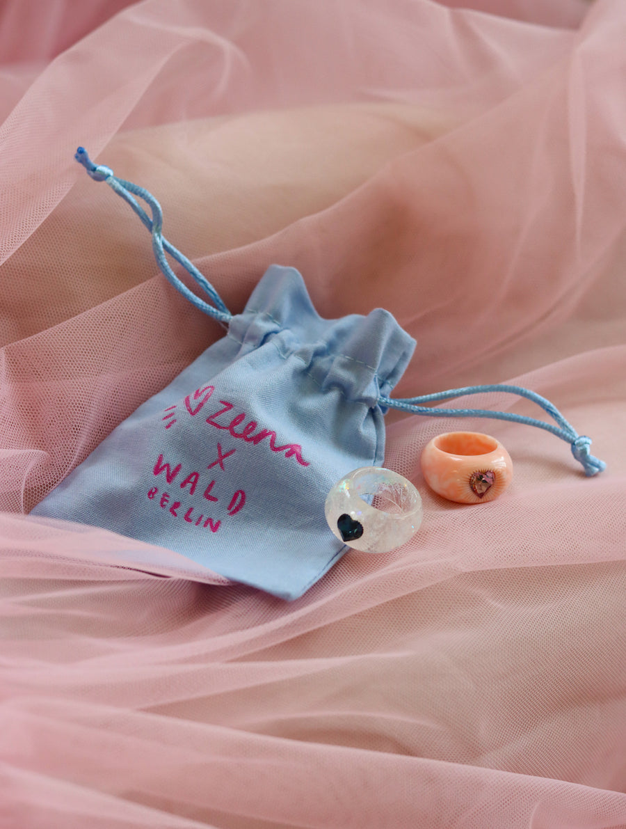 A light blue drawstring pouch labeled "Zeena x WALD Berlin" holds a SAMPLE Zeena x WALD Blue Heart Ring by WALD Berlin, beautifully displayed on pink tulle fabric.