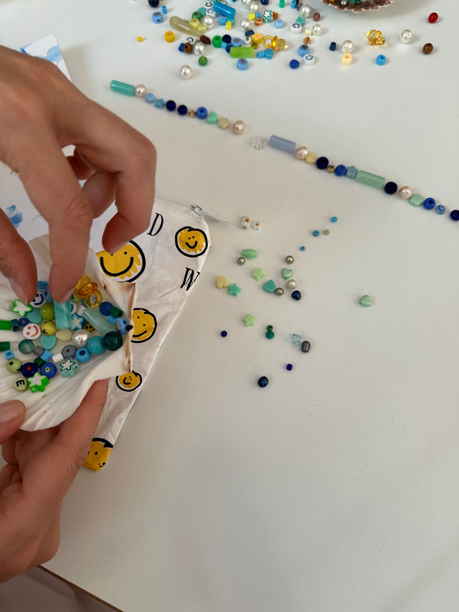 Hands arranging colorful beads on a white surface, with scattered beads and partially strung beadwork visible.