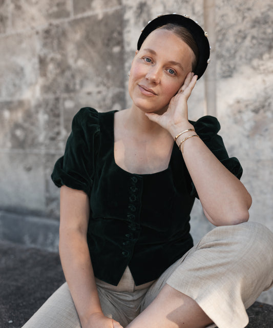 A woman in a dark green top and beige pants sits by a stone wall, gazing at the camera. She accessorizes with gold bracelets and the SAMPLE SALE Simone de Beauvoir Black Headband by WALD Berlin for a chic, relaxed look.