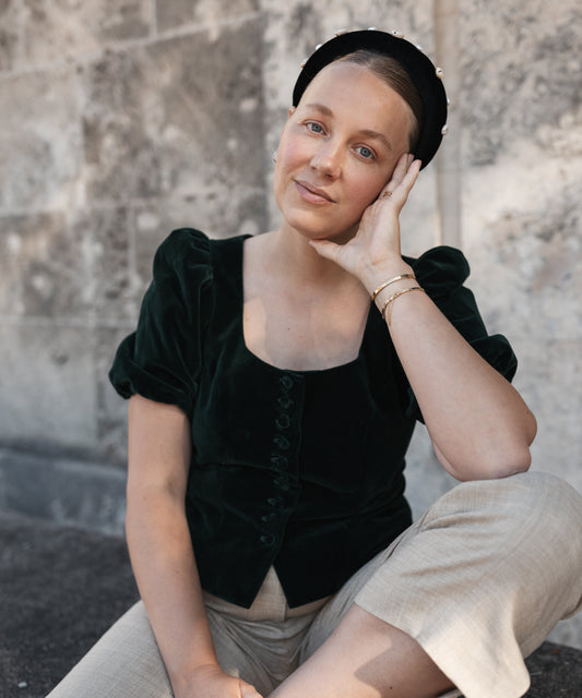 A woman in a dark green top and beige pants sits by a stone wall, gazing at the camera. She accessorizes with gold bracelets and the SAMPLE SALE Simone de Beauvoir Black Headband by WALD Berlin for a chic, relaxed look.