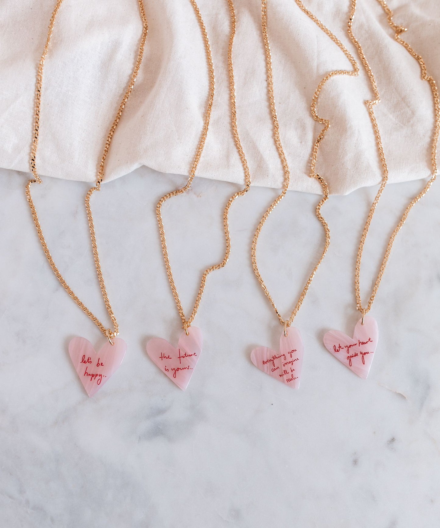 The WALD Berlin Let's be happy gold necklaces feature pink heart pendants with motivational messages, elegantly resting on a marble surface against a white polyester backdrop. Made in Germany, each piece radiates unique charm and craftsmanship.