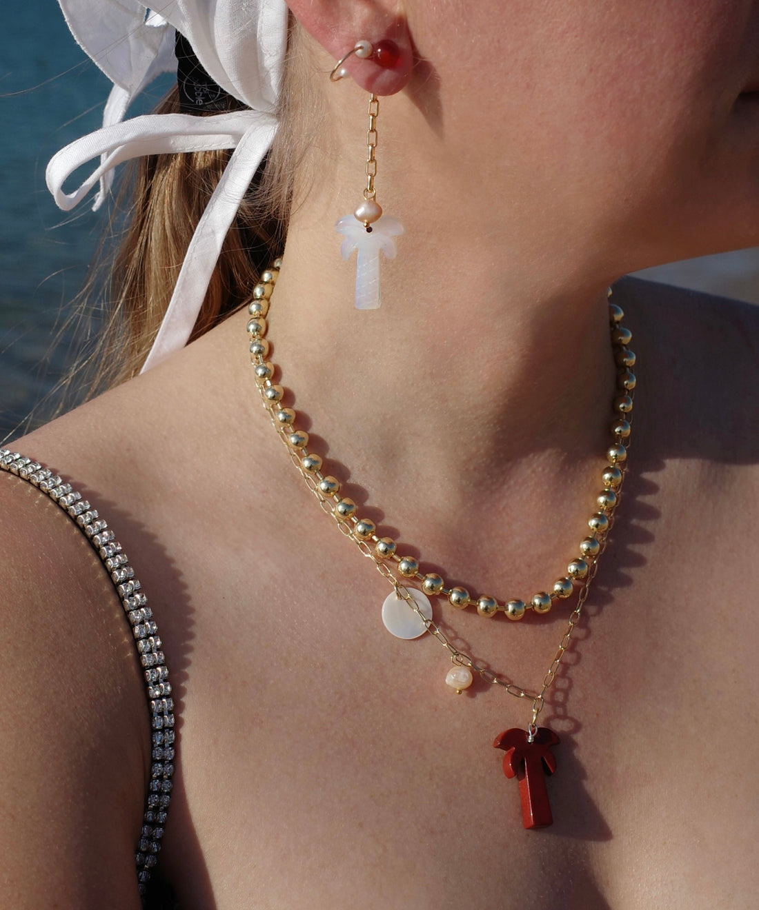 A woman stands near water wearing a white headscarf, layered gold necklaces with pendants, and WALD Berlin's Mexicana Mariachi Earrings Pearl.