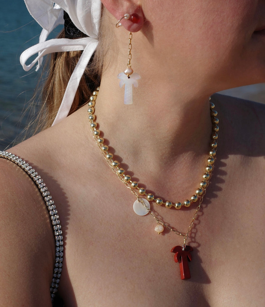 A woman stands near water wearing a white headscarf, layered gold necklaces with pendants, and WALD Berlin's Mexicana Mariachi Earrings Pearl.