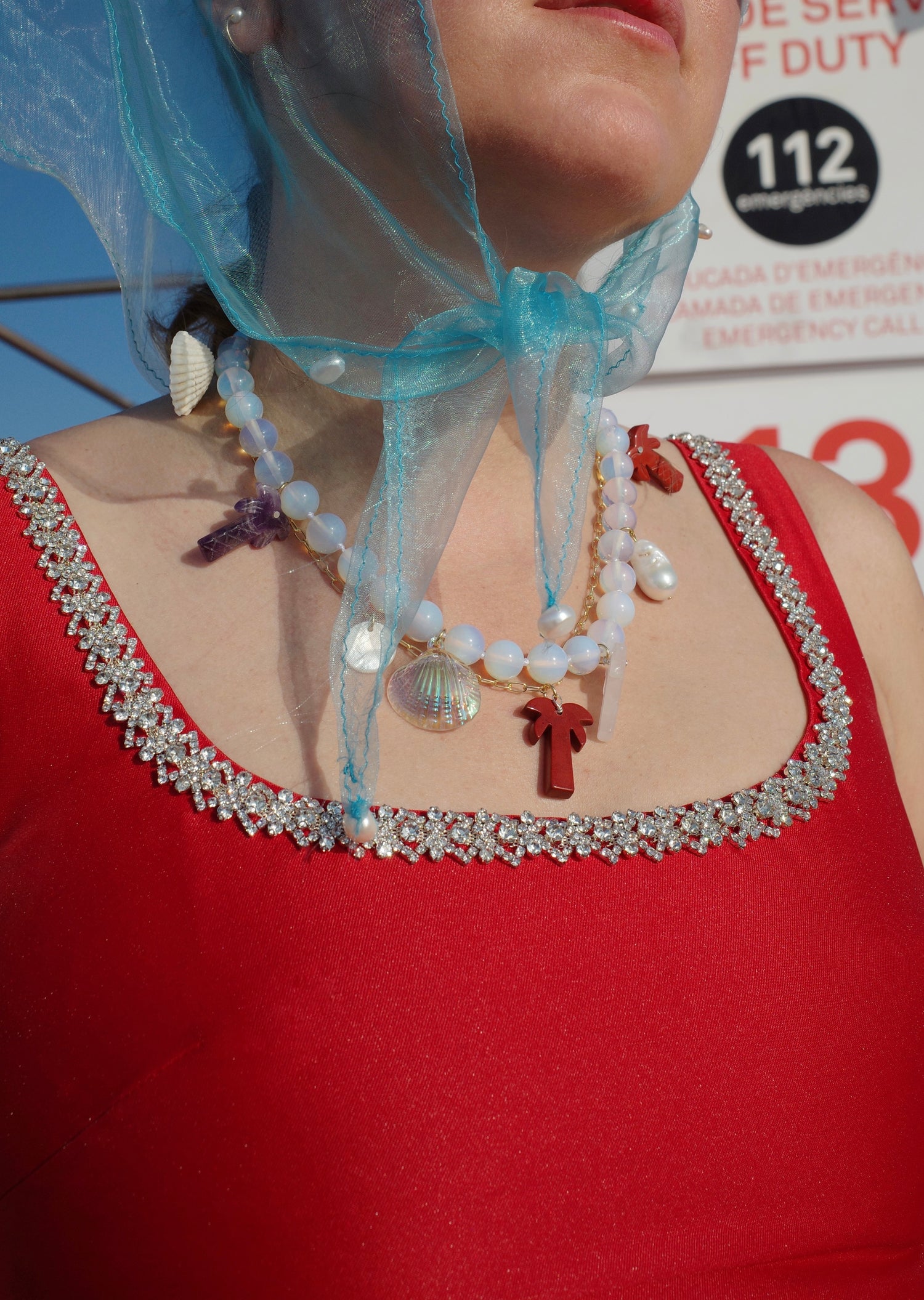 A person in a red dress with a jeweled neckline and layered necklaces—one is the Mexicana La Bamba Necklace by WALD Berlin—wears a blue sheer scarf tied under the chin. An emergency sign appears in the background.
