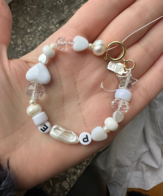A hand displays the WALD Berlin Personalized bracelet, crafted with a charming mix of white, pearl, and clear beads—including heart-shaped accents—and round letter beads spelling “PE.” Enhanced with sweetwater pearls and completed by a gold clasp, this custom piece radiates elegance and individuality.