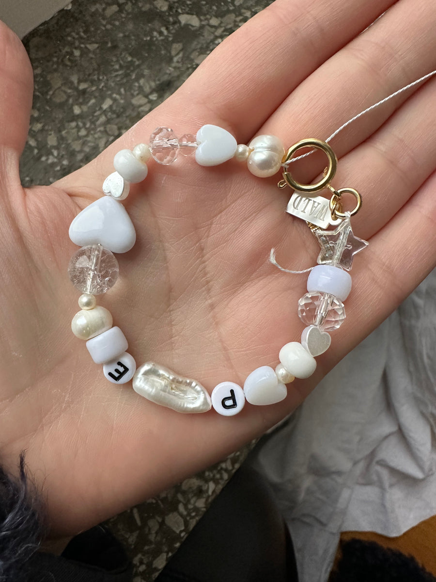 A hand displays the WALD Berlin Personalized bracelet, crafted with a charming mix of white, pearl, and clear beads—including heart-shaped accents—and round letter beads spelling “PE.” Enhanced with sweetwater pearls and completed by a gold clasp, this custom piece radiates elegance and individuality.