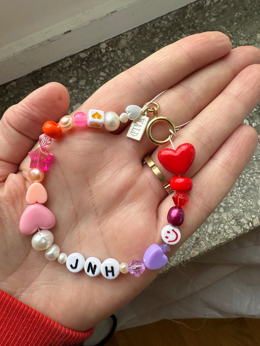 A hand holds the WALD Berlin Personalized bracelet, featuring colorful beads, sweetwater pearls, heart-shaped accents, a red heart charm, and custom letter beads spelling “JNH.”.