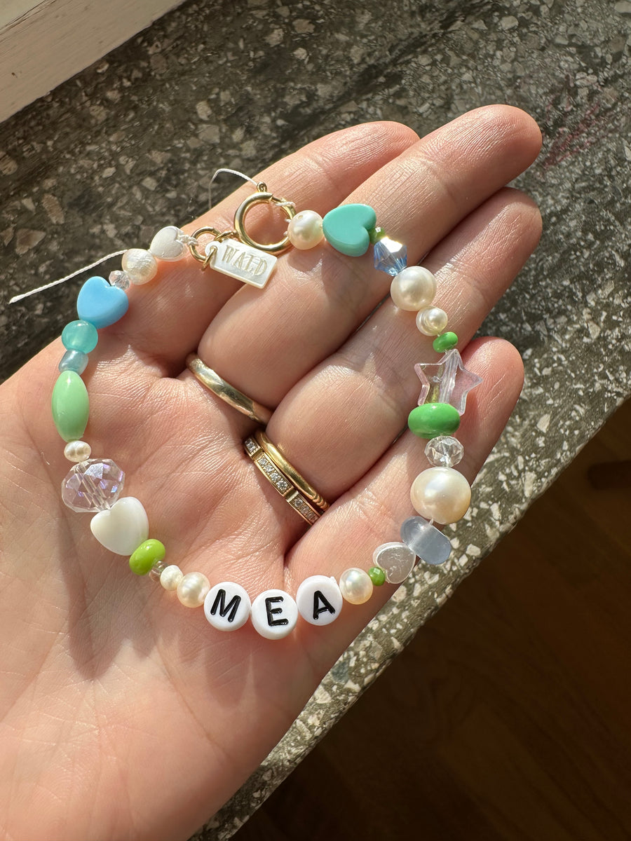A hand holds a WALD Berlin personalized bracelet featuring the letters “MEA,” heart and star-shaped beads, sweetwater pearls, green accents, and a clasp adorned with a small charm.