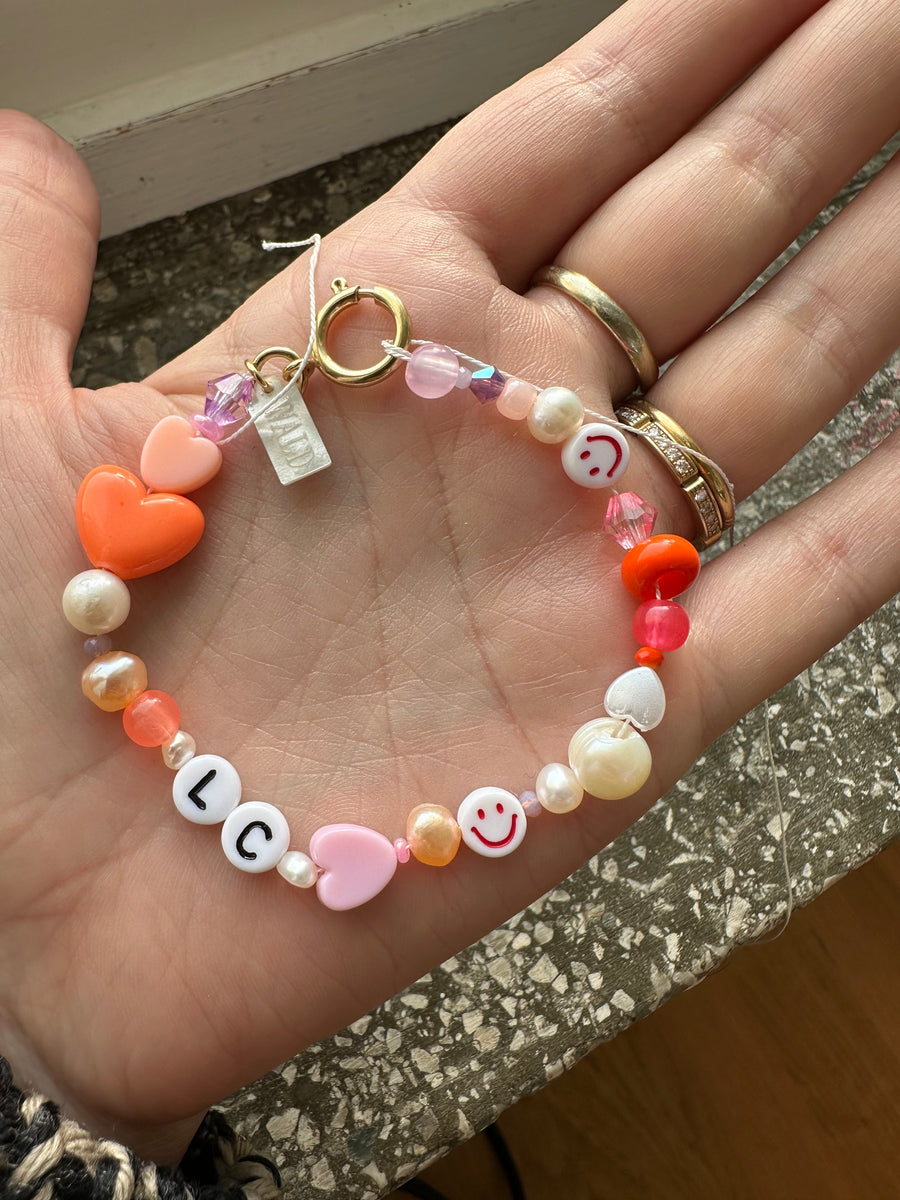 A hand holding the WALD Berlin Personalized bracelet, featuring colorful beads with hearts, sweetwater pearls, smiley faces, and the letters “L” and “C.”.