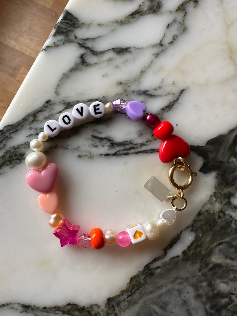 Discover the WALD Berlin Personalized Bracelet, featuring vibrant beaded hearts, stars, sweetwater pearls, and letter beads spelling “LOVE”—displayed on a marble surface. This charming accessory makes an ideal custom name or personalized bracelet for someone special.