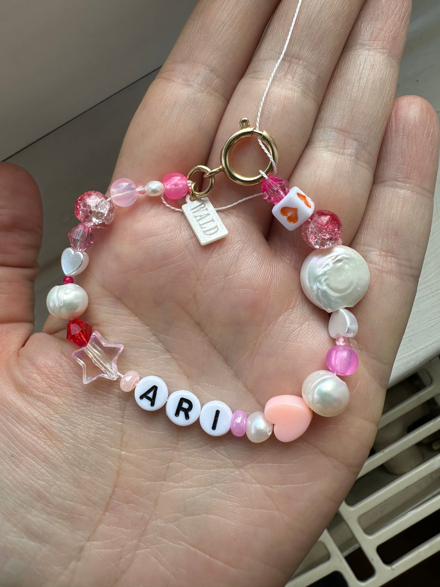 A hand holds the WALD Berlin personalized bracelet, featuring pink, white, and red beads with sweetwater pearls, a gold clasp, a “WILD” charm, and white letter beads spelling “ARI.”.