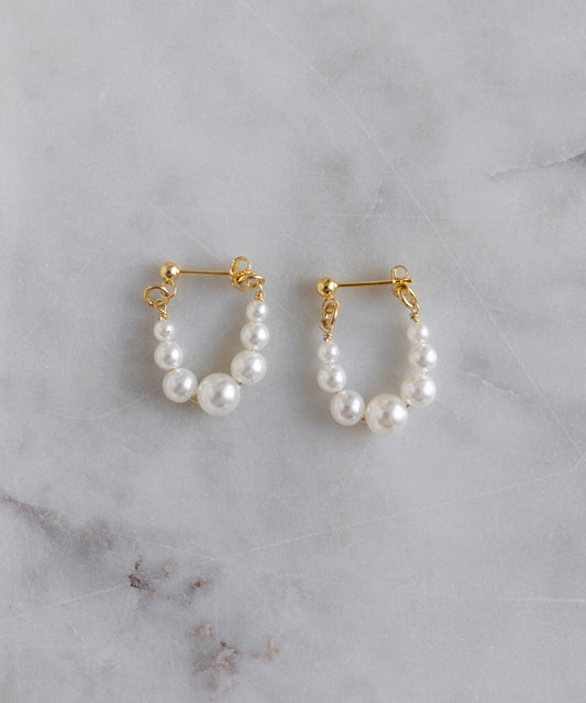 The Isla Bonita Pearl Hoops by WALD Berlin are gold plated earrings, each adorned with three sweetwater pearls, shown on a light gray marble surface.