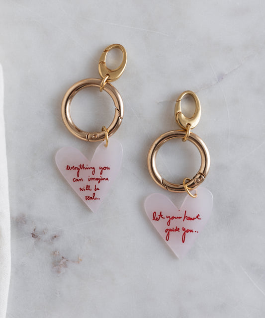 Two gold WALD Berlin key chains from the SAMPLE SALE, crafted in Germany from brass and bio polyester, each with a translucent heart pendant—one says "everything you can imagine will be real," the other, "let your heart guide you.