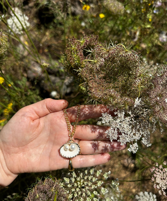 A hand holds the WALD Berlin Drop It Like It’s Hot Shell Necklace, featuring a shell and two pearls, against a backdrop of blooming wildflowers and Queen Anne's lace outdoors.