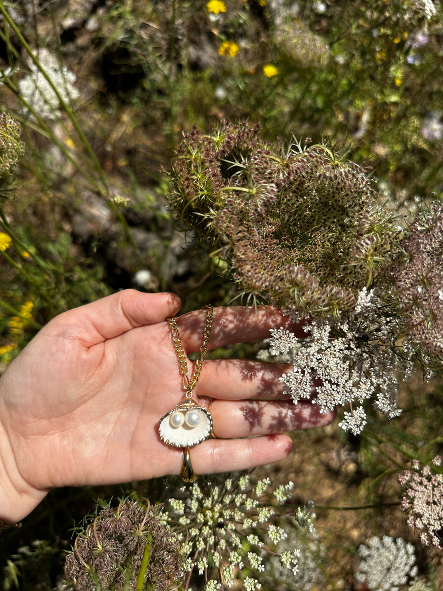 A hand holds the WALD Berlin Drop It Like It’s Hot Shell Necklace, featuring a shell and two pearls, against a backdrop of blooming wildflowers and Queen Anne's lace outdoors.