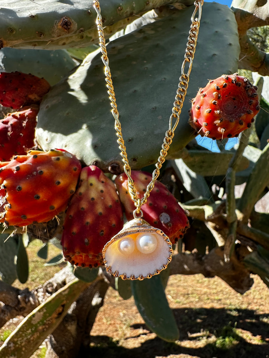 The Drop It Like It’s Hot Shell Necklace by WALD Berlin features a gold-plated chain, shell pendant, and two pearls, shown hanging before a prickly pear cactus with red fruit—a striking piece of handmade jewelry.