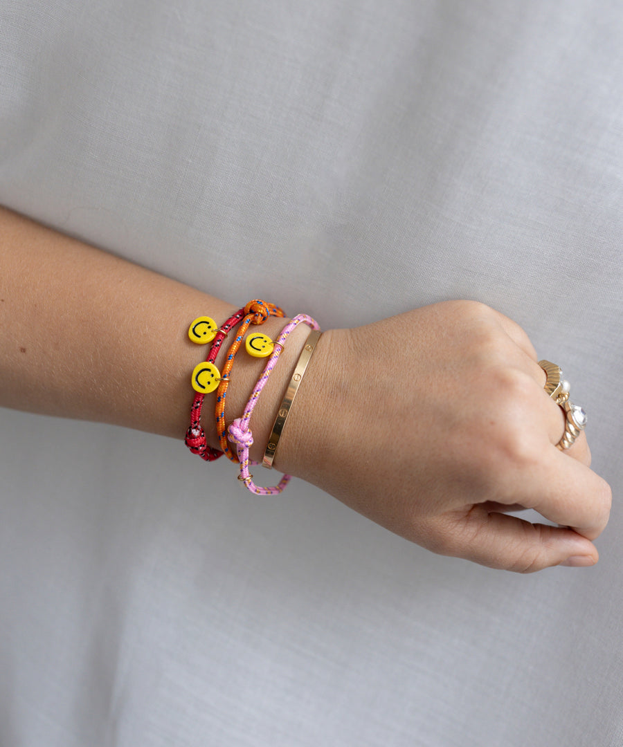 A wrist adorned with two braided bracelets featuring smiley face beads and a gold metal bracelet includes the Smilie Dude Pink Friendship Bracelet by WALD Berlin.