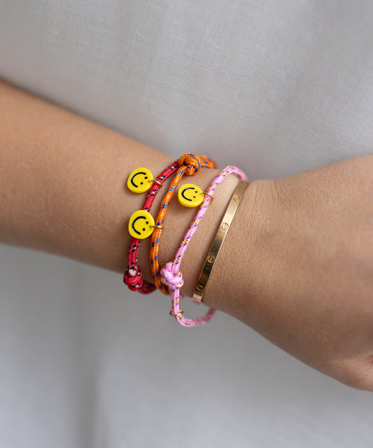 A wrist adorned with WALD Berlin's Smilie Dude Orange Friendship Bracelet alongside two other colorful braided string bracelets featuring smiley face beads, paired with a single gold metal bracelet.