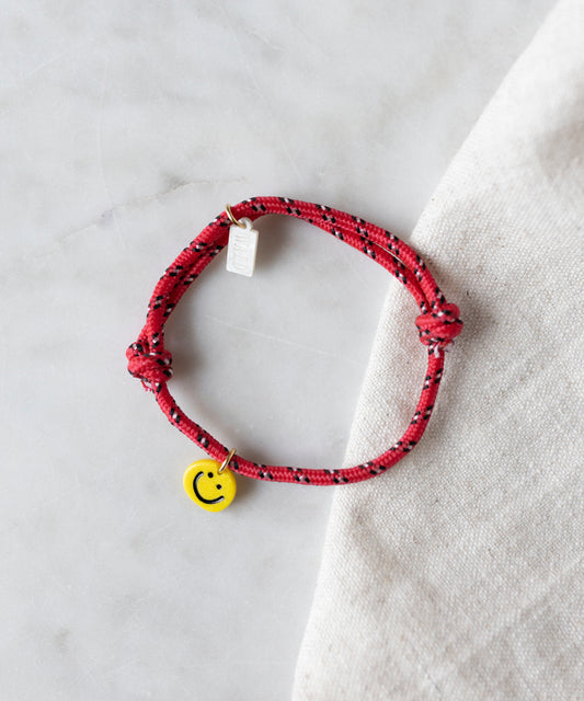 The Smilie Dude Red Friendship Bracelet by WALD Berlin, featuring a yellow smiley face charm, is displayed on a marble surface next to light-colored fabric.