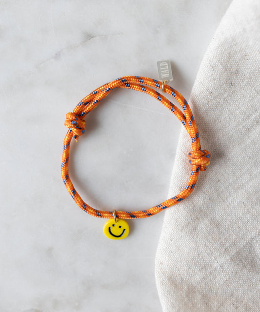 The WALD Berlin Smilie Dude Orange Friendship Bracelet, featuring an orange, blue, and black woven pattern with a yellow smiley face charm, rests on a light fabric surface.