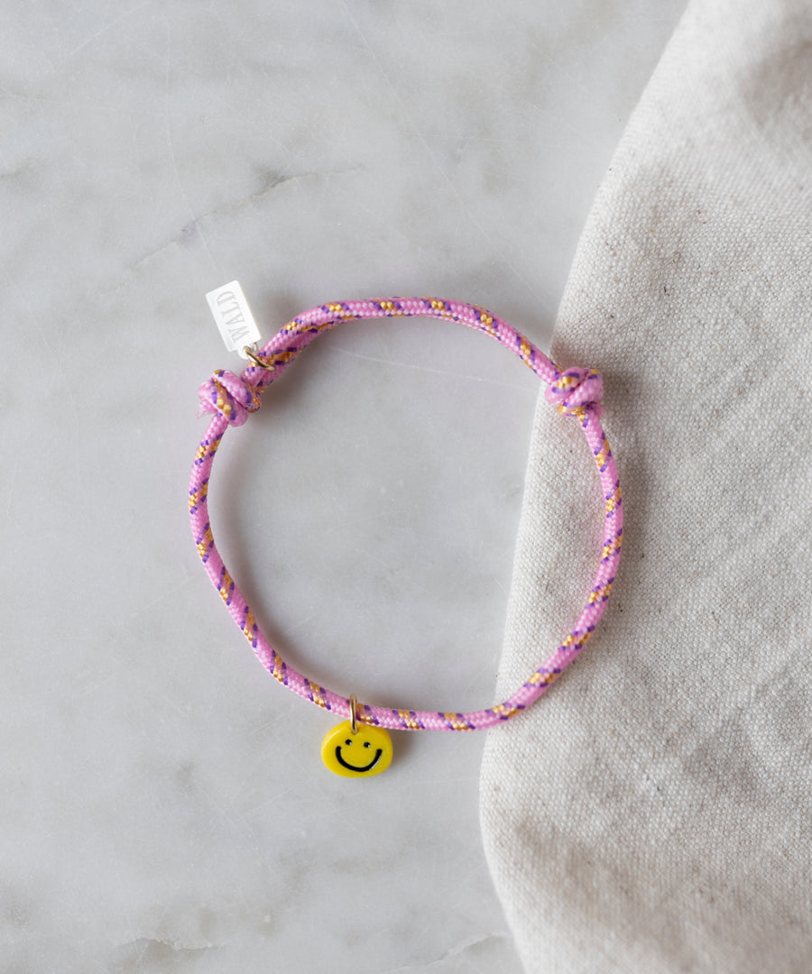 The Smilie Dude Pink Friendship Bracelet by WALD Berlin, featuring a yellow smiley face charm, is displayed on a marble surface.