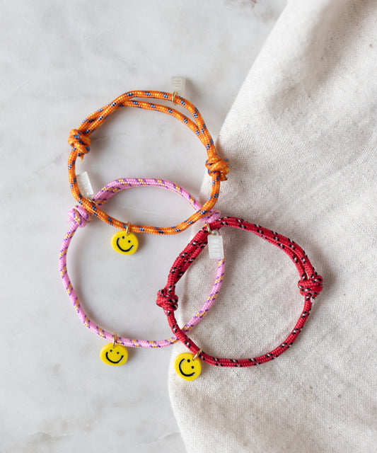 The WALD Berlin Smilie Dude Orange Friendship Bracelet, along with red and pink woven rope bracelets featuring small yellow smiley face charms, are displayed on a light fabric background.