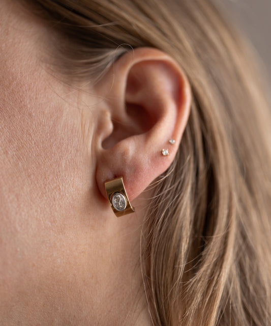Close-up of a person's ear showcasing three piercings. The main piercing features WALD Berlin's Isla Bonita just a friend earring, a unique gold piece with a round gemstone made from recycled gold, while the upper ear has two small stud earrings.
