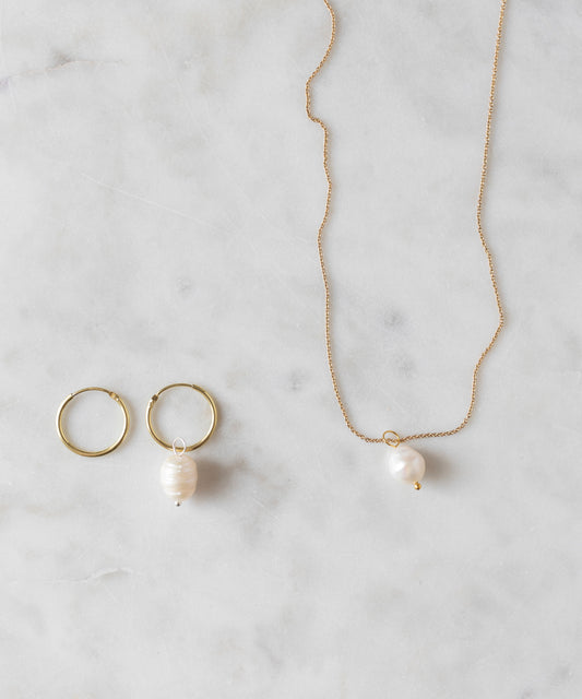 Presenting the Sweetwater Pearl Charm Set by WALD World: gold hoop earrings adorned with sweetwater pearl charms, paired with a coordinating gold chain necklace featuring a matching pearl pendant—elegantly displayed on a marble surface.
