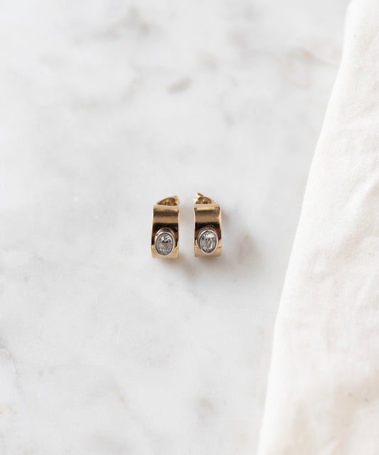 The Isla Bonita just a friend earrings by WALD Berlin, delicate stud earrings crafted from recycled gold and adorned with clear gemstones, are elegantly displayed on a marble surface beside a white cloth.