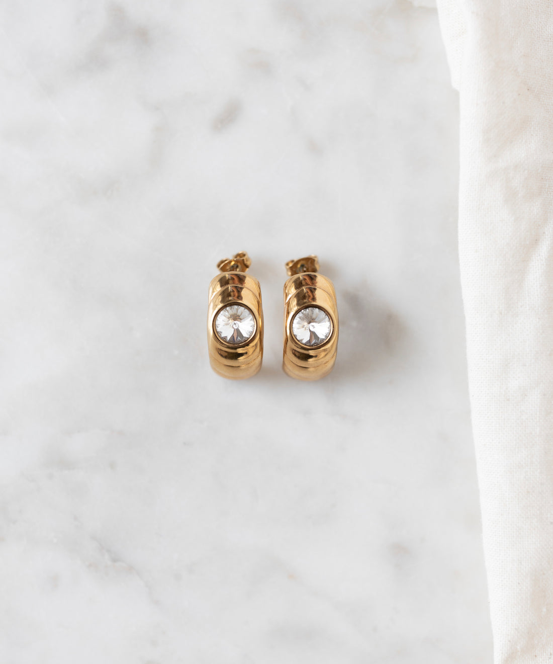 The Isla Bonita Ready or Not earrings by WALD Berlin, featuring a single clear Swarovski stone on each gold hoop, rest elegantly on a marble surface beside a white fabric, capturing an 80s vibe.