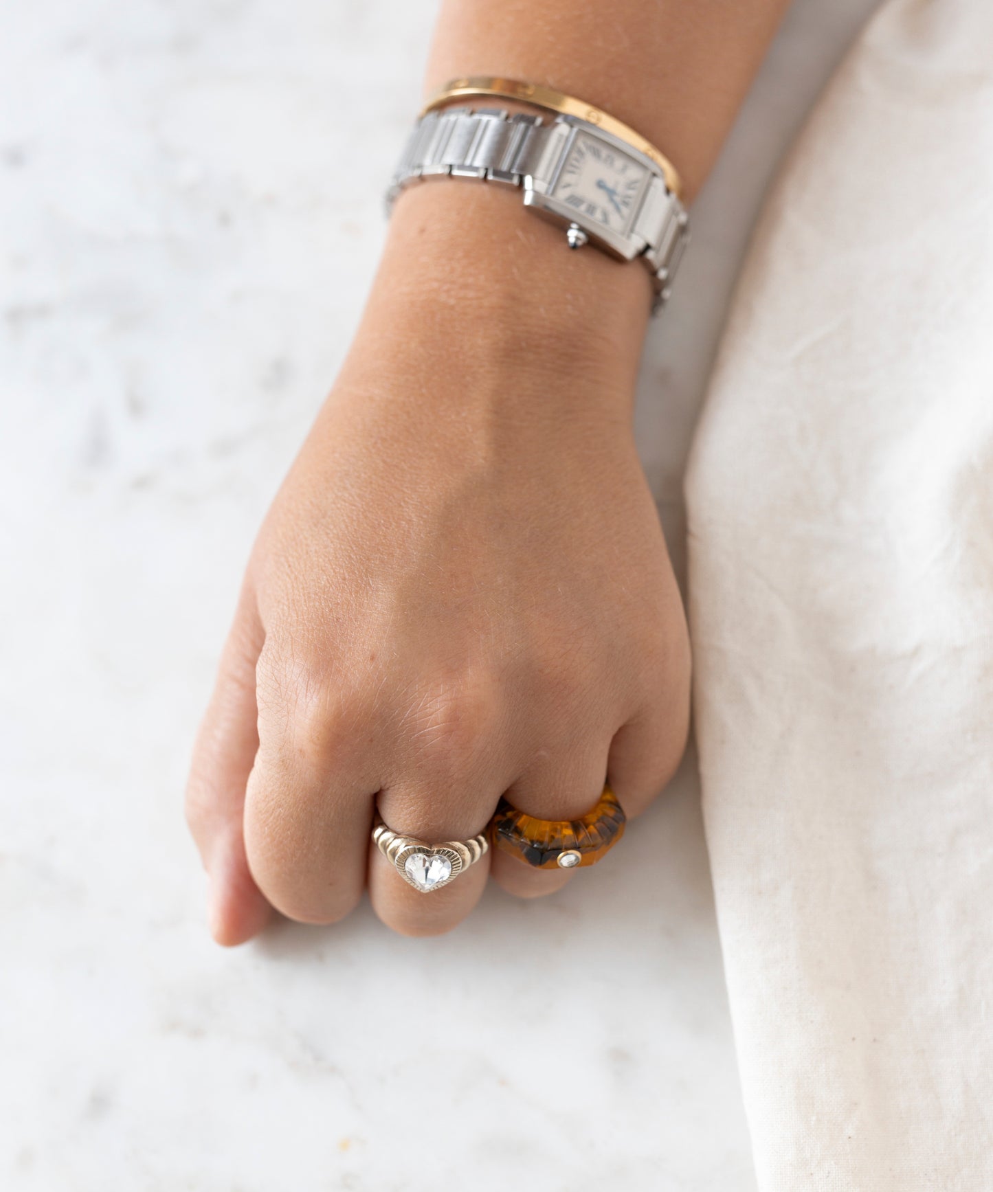 A close-up of a person's hand wearing two rings, including the Isla Bonita Shining Star ring by WALD Berlin, and a silver wristwatch with a white face, resting on an amber-colored marble surface next to a white fabric.