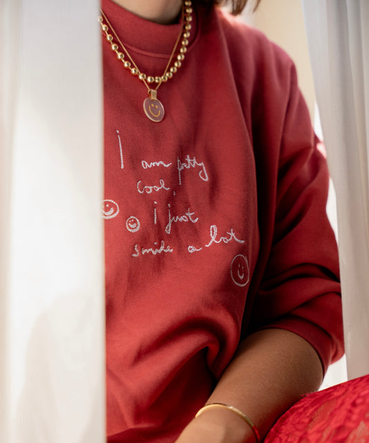 A person is wearing a PRETTY COOL Jumper by WALD Berlin, crafted from bio cotton in a limited edition Bordeaux color, paired with a gold necklace. Only part of their face is visible.