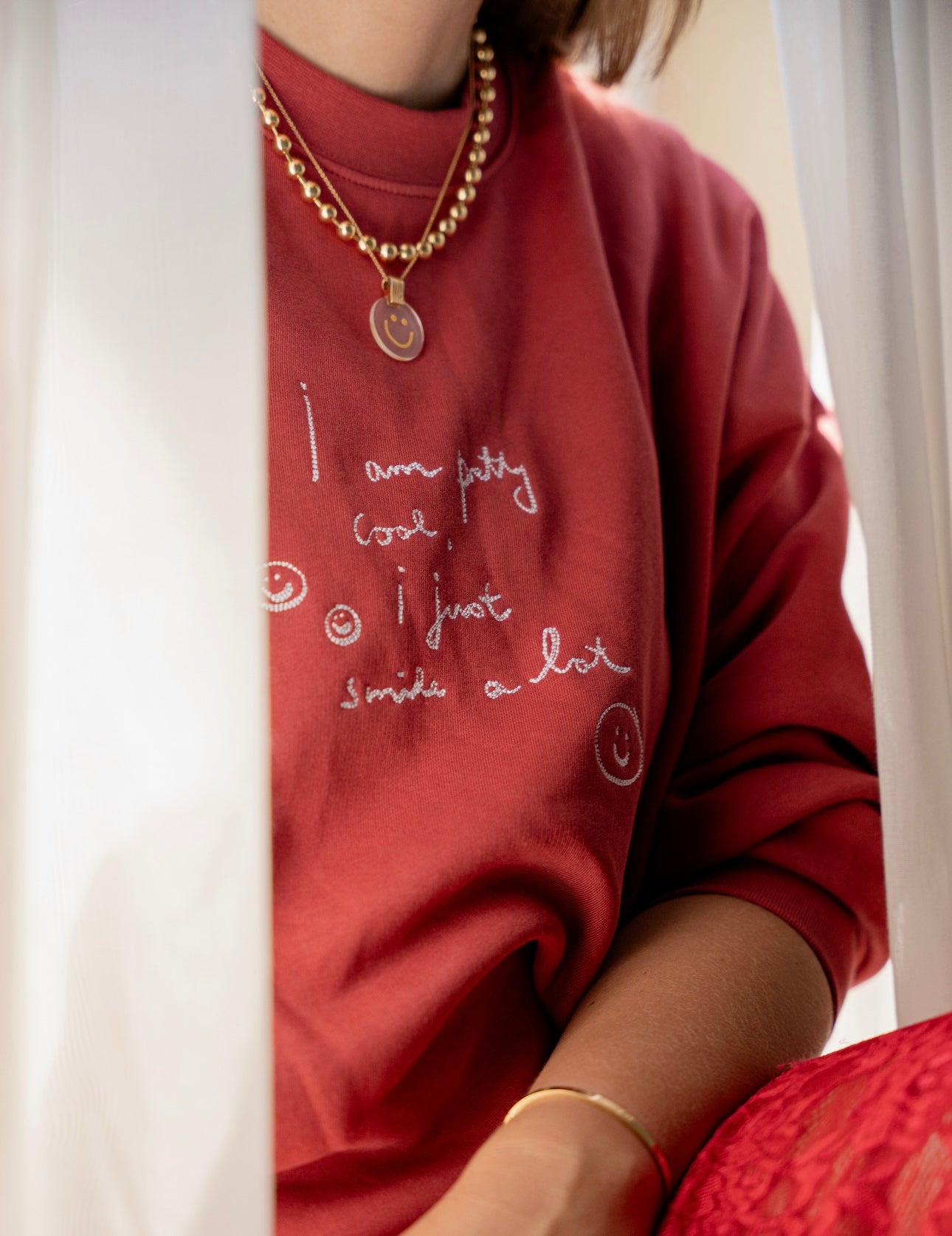 A person is wearing a PRETTY COOL Jumper by WALD Berlin, crafted from bio cotton in a limited edition Bordeaux color, paired with a gold necklace. Only part of their face is visible.