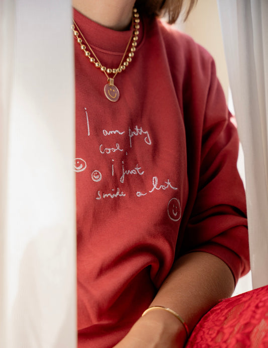 A person is wearing a PRETTY COOL Jumper by WALD Berlin, crafted from bio cotton in a limited edition Bordeaux color, paired with a gold necklace. Only part of their face is visible.