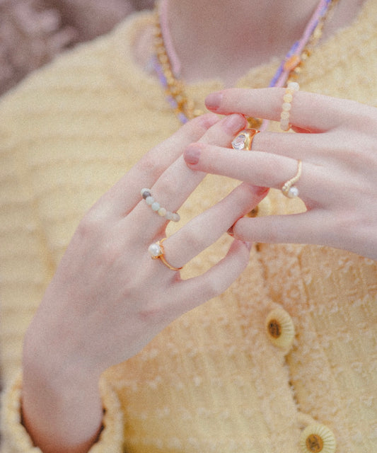 A person wearing a textured yellow jacket and multiple beaded rings, including the WALD Berlin SAMPLE SALE Pop Pearl Ring Mini, holds their hands together.