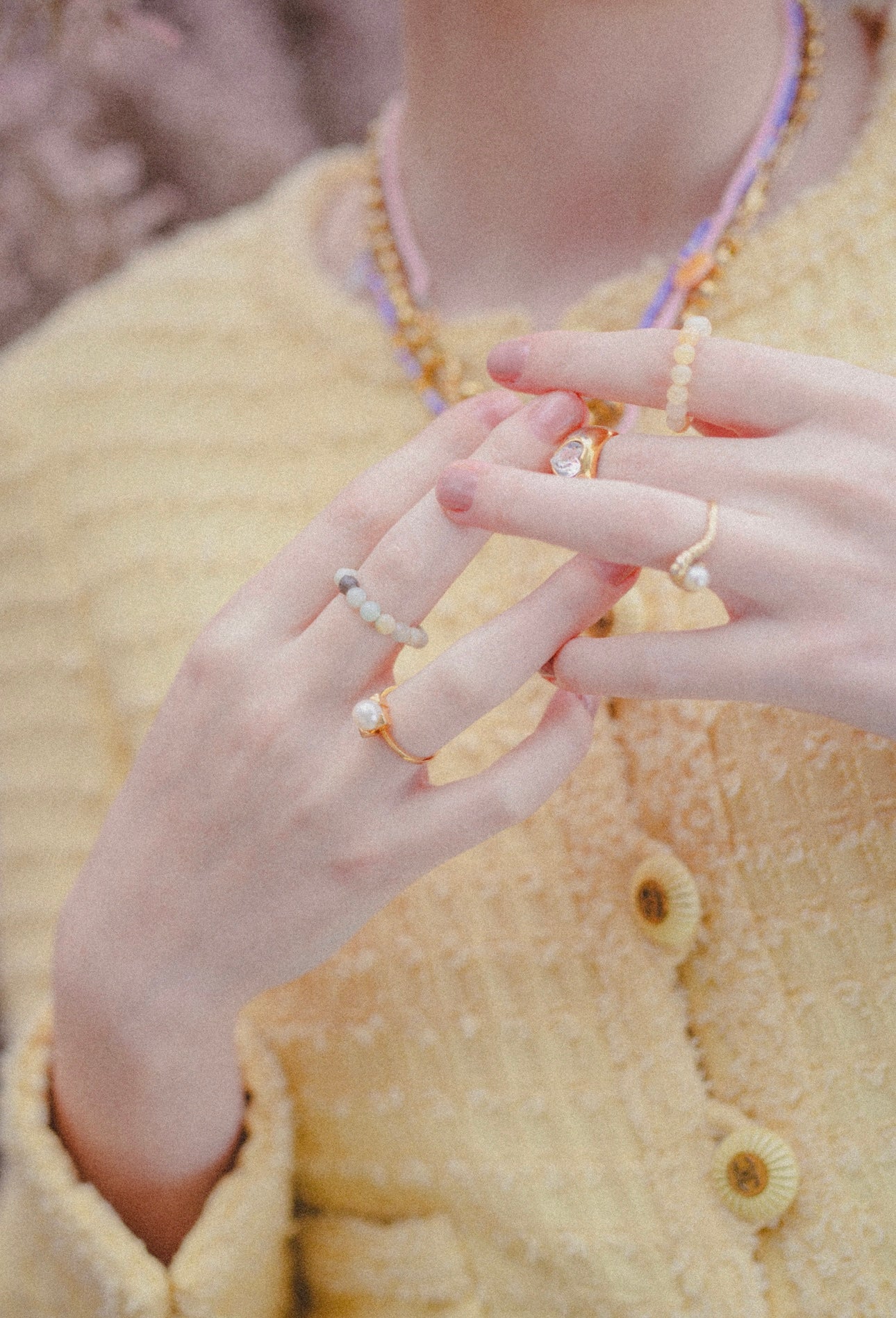 A person wearing a textured yellow jacket and multiple beaded rings, including the WALD Berlin SAMPLE SALE Pop Pearl Ring Mini, holds their hands together.